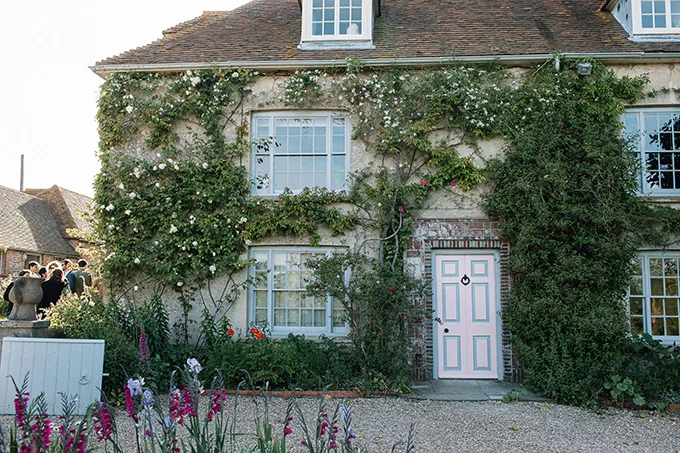 A country house with a pale pink front door and climbing plants growing up the front. 