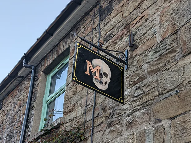 A black shop sign with a skull and a letter M on it, hanging from a stone wall. 
