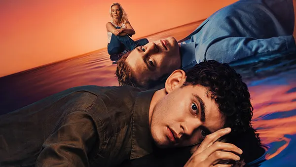 2 young white men lying down on a stylised bed of sand, with a white woman sitting upright in the background. 