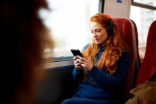 A young white woman with long red hair sitting next to a window on a train, wearing headphones and looking at her mobile phone. 