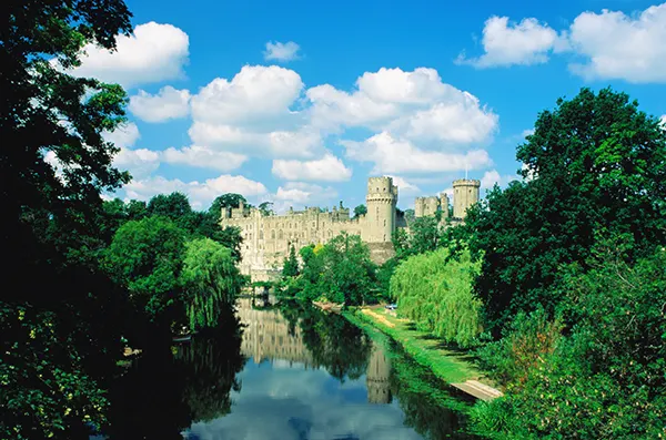 An old English light stone castle with towers and battlements, set in woodlands next to a river.