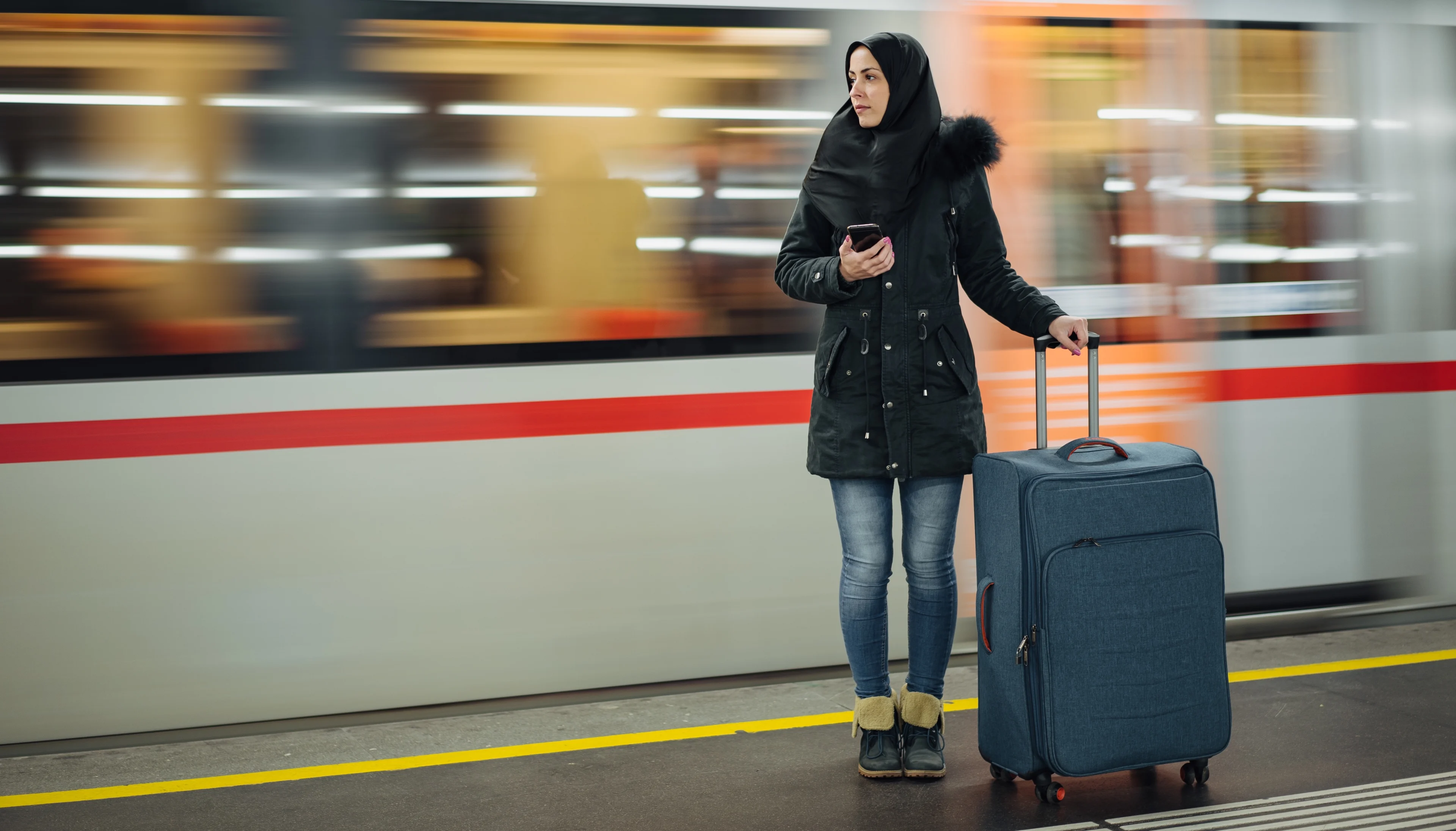 A Muslim woman with a large suitcase on a train platform as a train goes by in the background