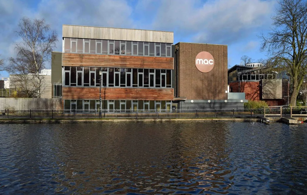A modern brick, wood and glass building next to a lake. A large red circular sign with white letters saying MAC is high up on a wall.