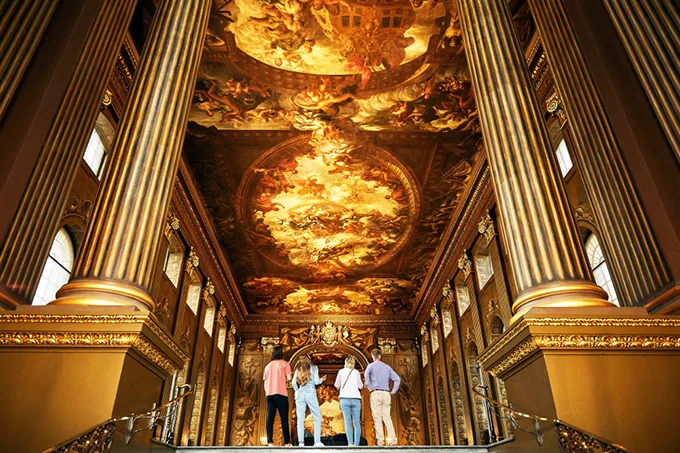 4 people with their back to the camera standing in a huge and ornately decorated hall with high columns and painted walls and ceiling.
