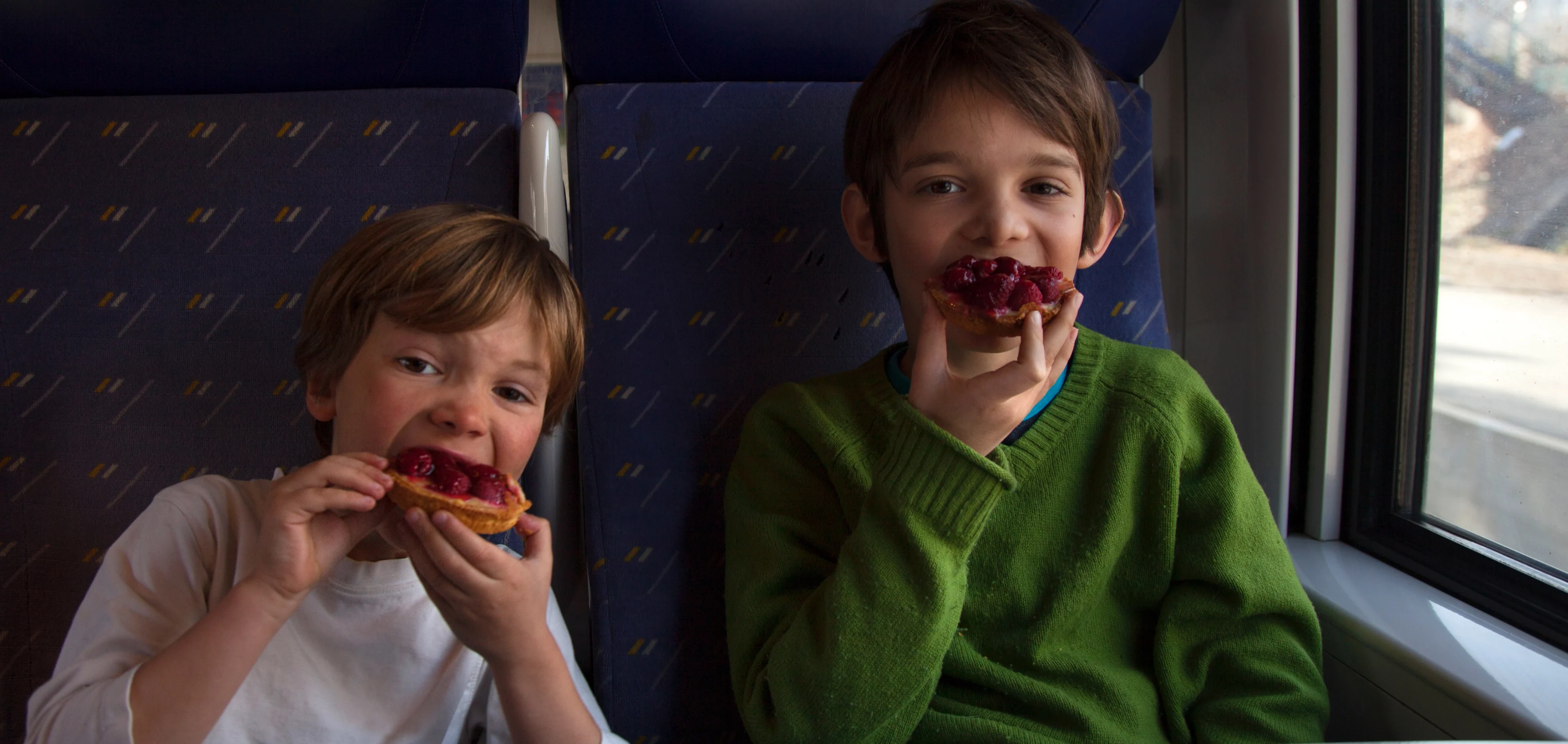 2 young boys eating large cakes while sitting at a table on a train