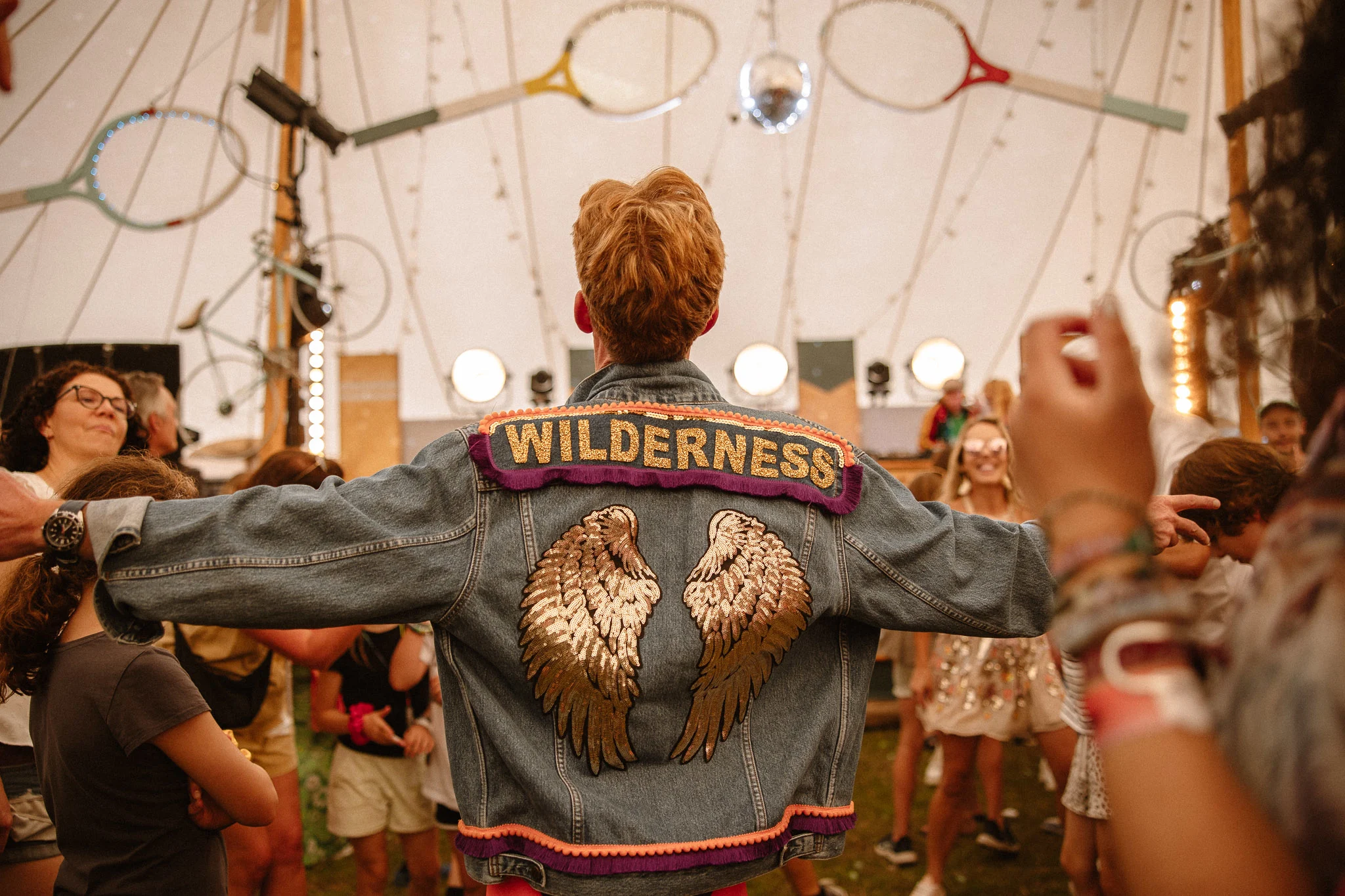 A person wearing a denim jacket embroidered with gold wings and "Wilderness" dances in a festival tent with hanging rackets.