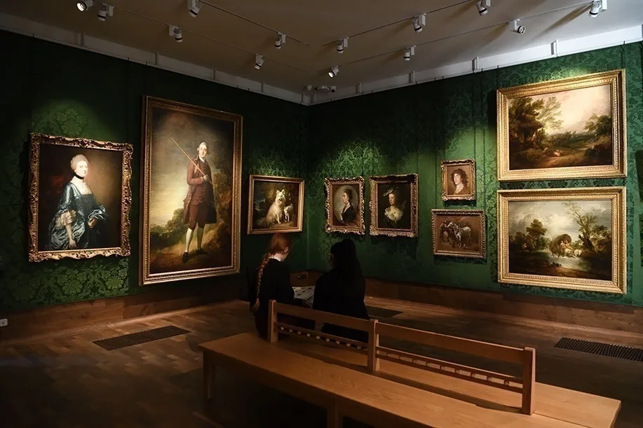 An art gallery with a number of portraits hung against green wallpapered walls. 2 people are sitting on a wooden bench looking at the art. 