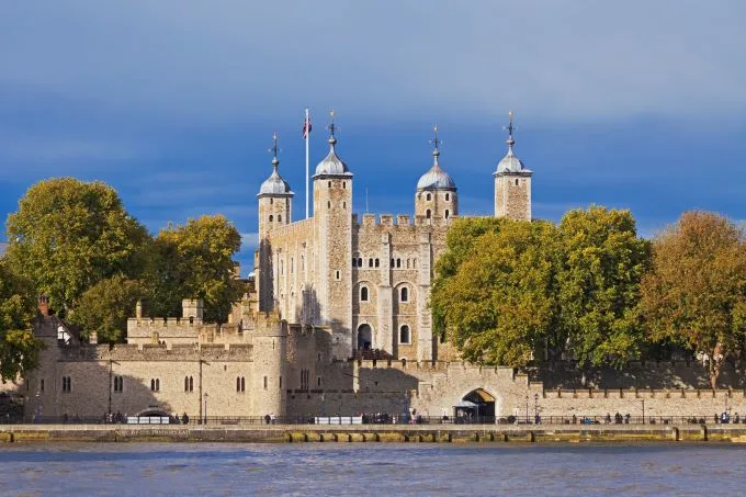 The Tower of London sits on the River Thames, surrounded by green trees and sitting under a hazy blue sky.
