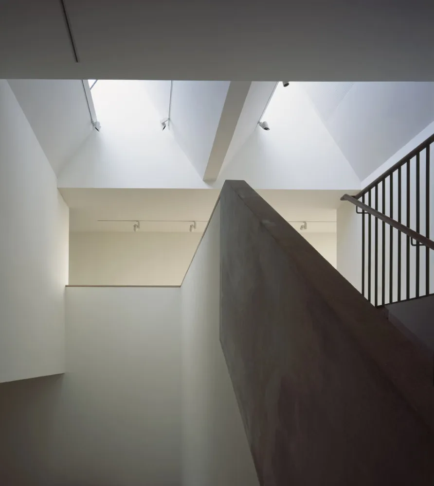 Light shines in from skylights down into a large gallery space with white walls and a concrete staircase. 