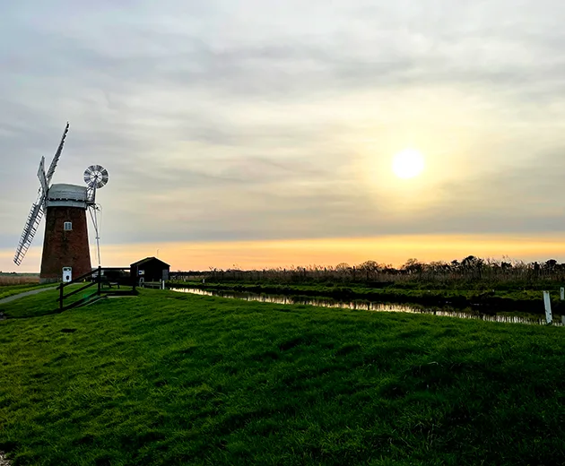 A windmill on a grassy bank next to a canal on the Norfolk Broads, with the sun low in the sky. 