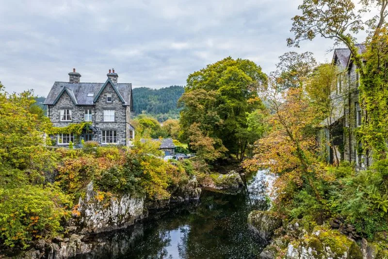 A stone covered two-story house sits on the banks of a river, surrounded by green trees, underneath a cloudy sky.