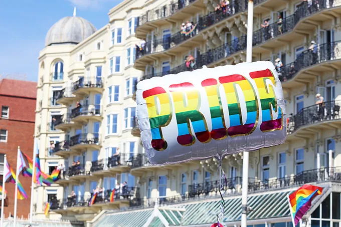 A helium balloon with the word PRIDE in rainbow letters, floating in front of white buildings with rainbow flags flying. 