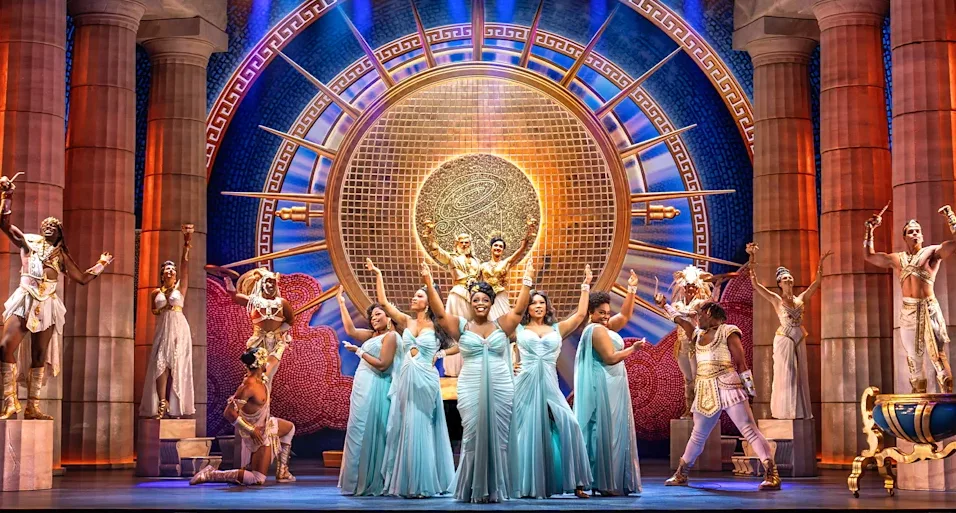 An ensemble cast on stage for Disney's Hercules, including 5 Black women in blue dresses centre stage and men in tunics standing on pillars either side, with a golden sun backdrop. 