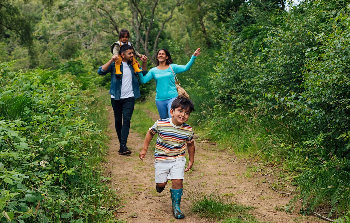 A happy smiling family of 4 are walking down a woodland path. A young boy in a t-shirt, shorts and wellington boots runs ahead while his mother and father walk behind. The father is carrying the young daughter on his shoulders. 