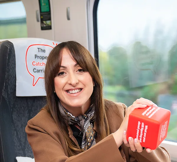 Actress Natalie Cassidy sitting at a train table and holding a red dice with conversation prompts on it, in "The Proper Catch-Up Carriage".