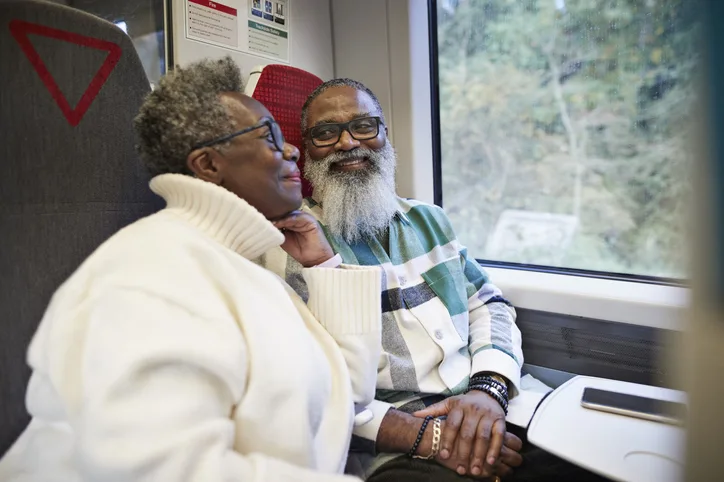 An older Black man and woman sit smiling looking out of a train window.