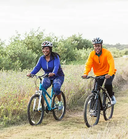 A smiling middle-aged Black woman and Black man in athletic clothing and cycle helmets riding bicycles along a grassy path in the countryside.