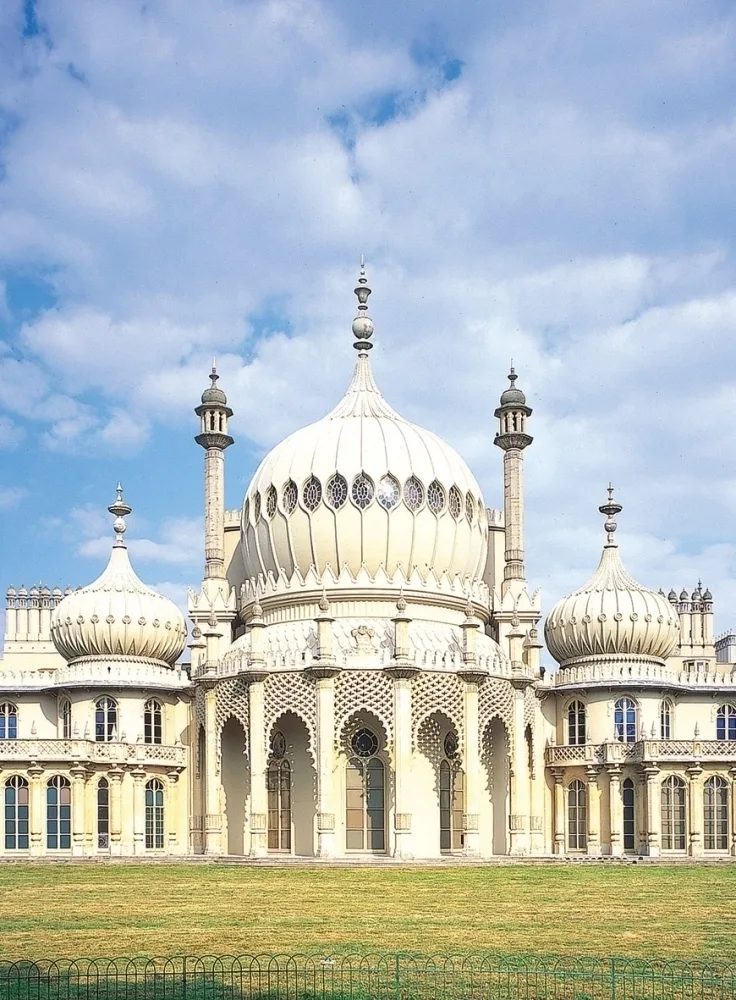 The Royal Pavilion in Brighton, a white building with multiple domes, turrets and columns, with a green lawn in front and blue sky with white clouds behind. 