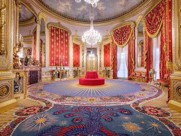 A lavishly appointed room in a stately home, with a large chandelier, a painted ceiling, red velvet draped curtains and brightly coloured carpet with a red circular seat in the centre of the room.