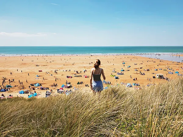 Rear view of a white girl looking out to sea over a busy, sandy beach on a sunny day. 