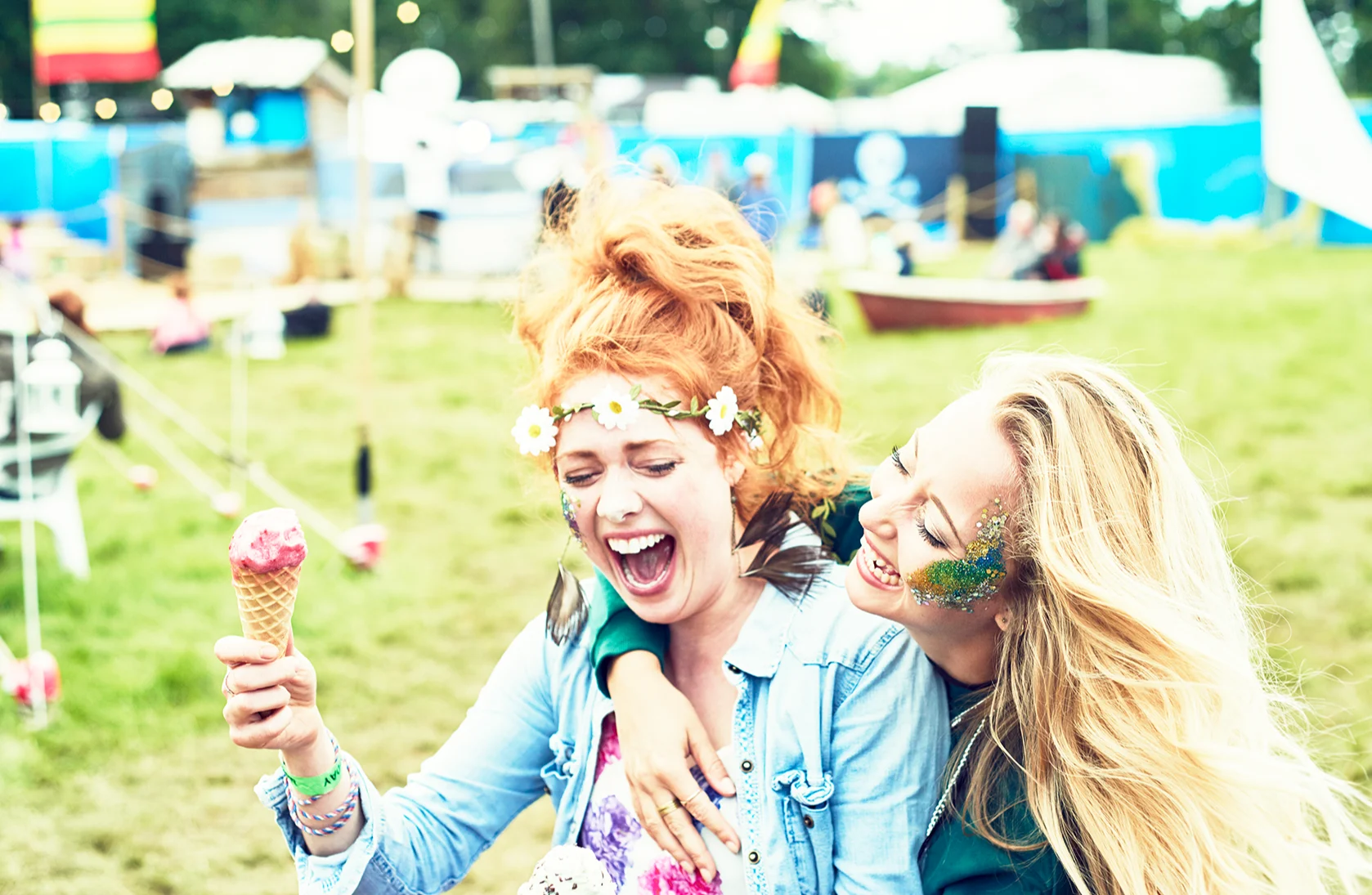 2 white young women sitting on the grass at a festival, they are laughing and one is holding an ice cream cone