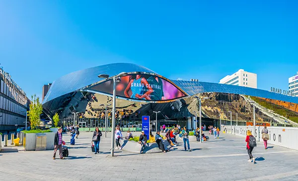 A modern station building with mirrored panels and curving surfaces, and a large video screen. People are walking about the paved area in front.