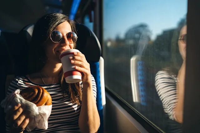 A happy young woman wearing sunglasses holds a large pastry and drinks a hot drink while looking out of a train window