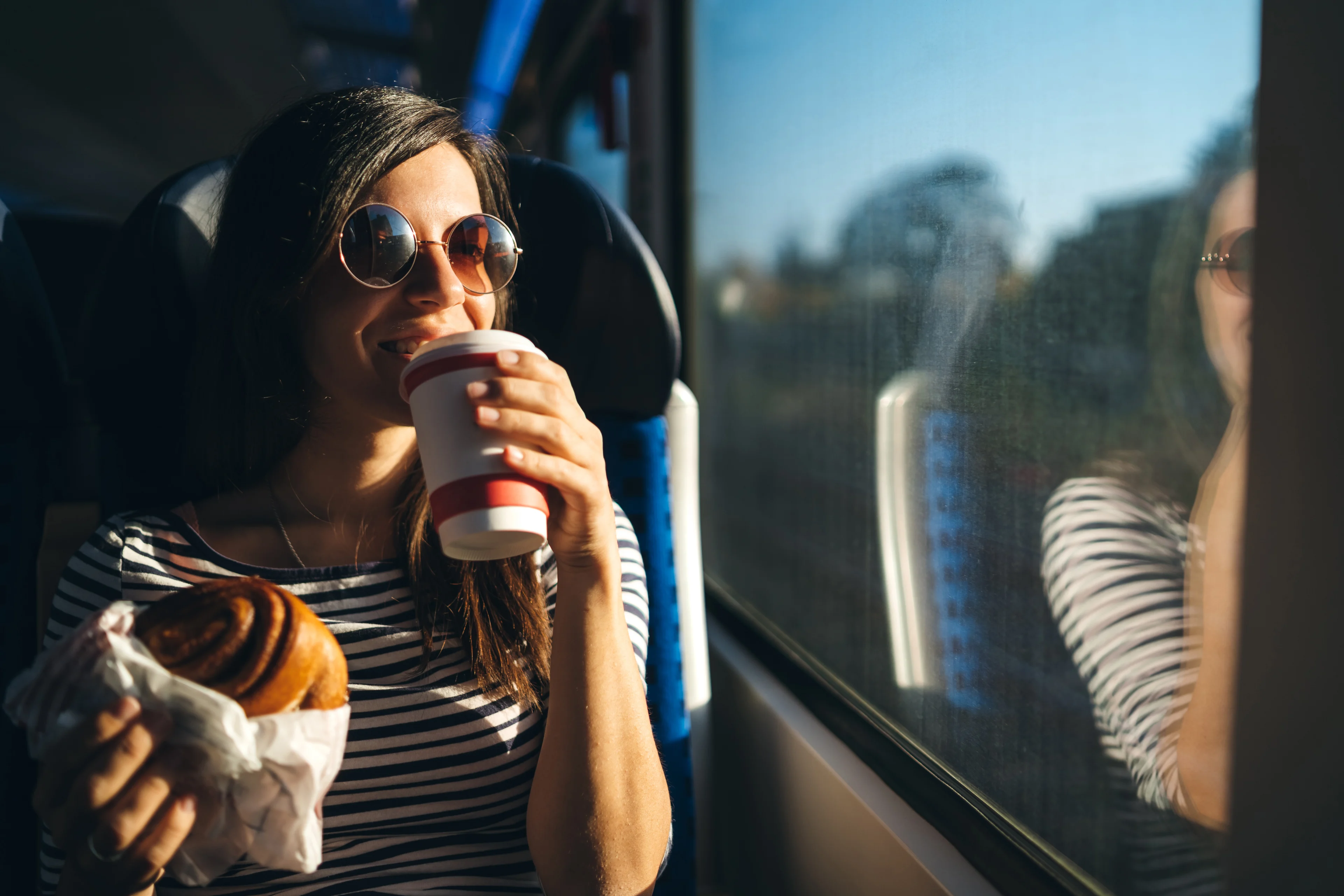 A happy young woman wearing sunglasses holds a large pastry and drinks a hot drink while looking out of a train window