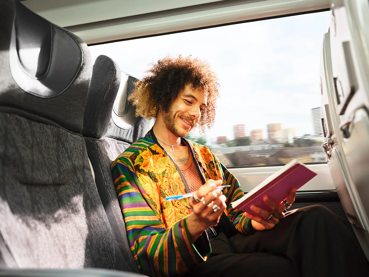 A Black man with curly hair and beard, wearing colourful patterned jacket using a tablet while seated on a train with view of a city from the window. 