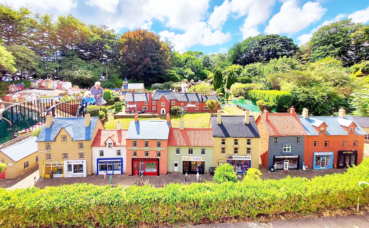 A detailed model village featuring colourful shops and houses, surrounded by lush greenery, with visitors viewing the display.