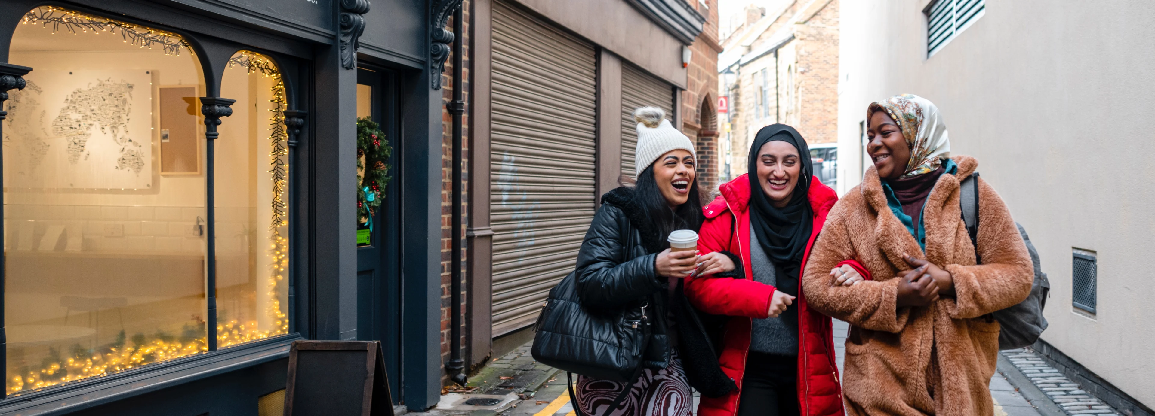 Three female friends, 1 is wearing a bobble hat and 2 have headscarves, are smiling and walking arm in arm down a narrow street