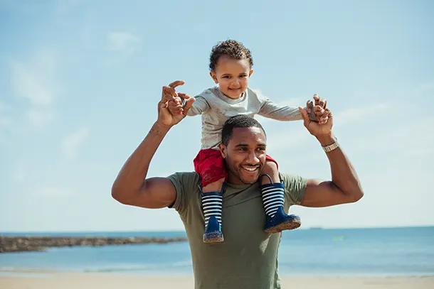 A smiling Black man walks along a beach holding a Black toddler on his shoulders