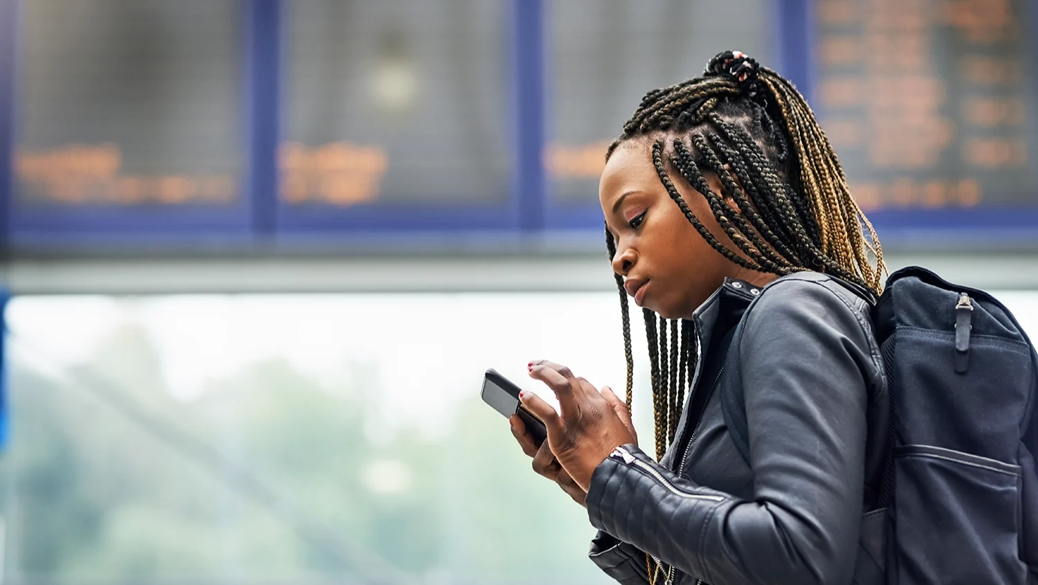 A young Black woman with braided hair checks her mobile phone at a railway station