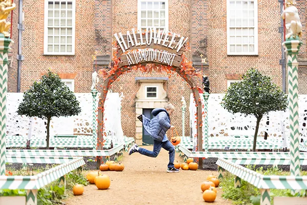 A white boy holding a pumpkin basket and jumping in front of a display with a "Happy Halloween" arch, orange pumpkins and green topiary trees outside a brick building..