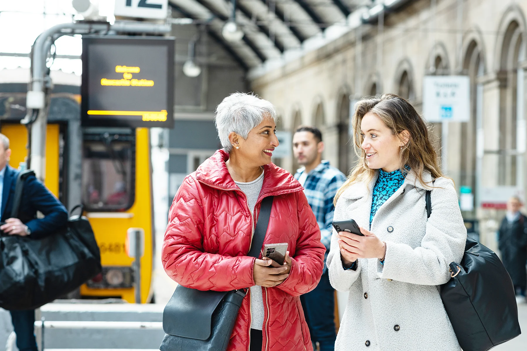 A middle-aged woman with short grey hair and a younger woman with long brown/blonde hair chat at a train station while checking their phones. 
