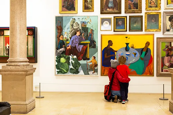 A woman kneeling with a child standing in front of a wall of paintings in a bright art gallery. 