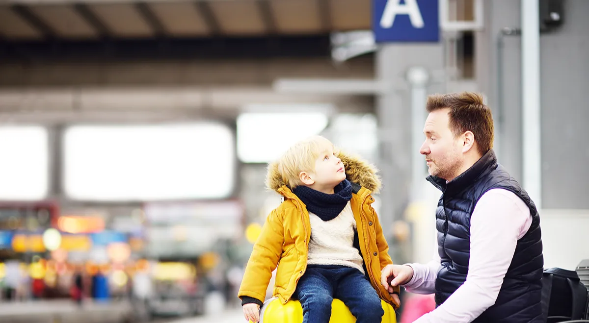 Child in yellow coat sitting on large yellow suitcase, talking with adult at train station platform.