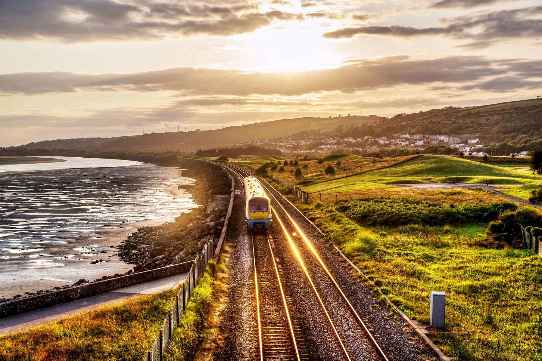 A train going along a coastal track at sunset, with the sea on one side and green fields and hills on the other. 