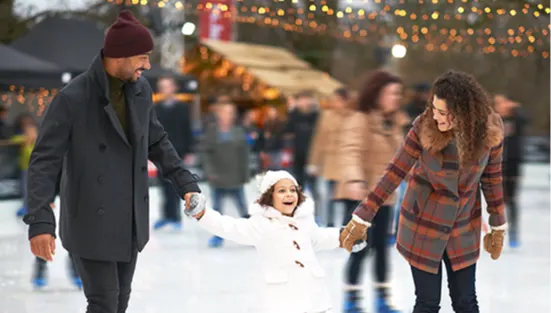 A father and mother holding the hands of a young girl as they skate on an outdoor ice rink. 