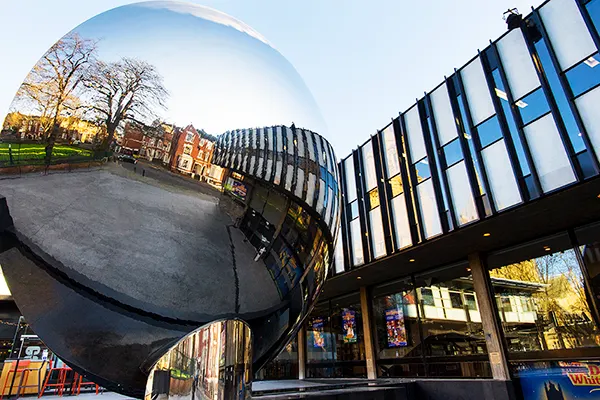 A huge mirror sculpture reflecting a modern building on concrete stilts. 