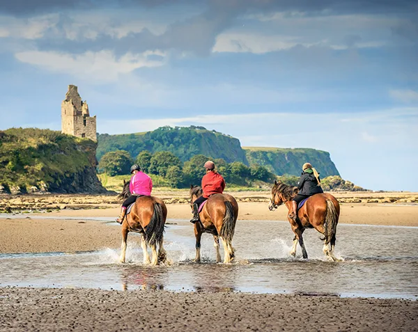 3 horse riders viewed from behind on a sandy beach with large pools, green hillside with the ruins of an old building is visible in the distance. 