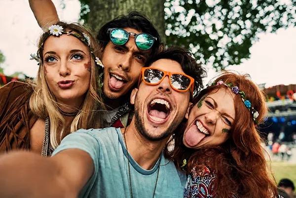 Four friends in festival attire pulling silly faces for a selfie outdoors, wearing flower crowns and colourful sunglasses.