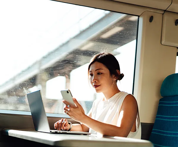 An East Asian woman sitting at a train table with an open laptop and looking at her phone. 