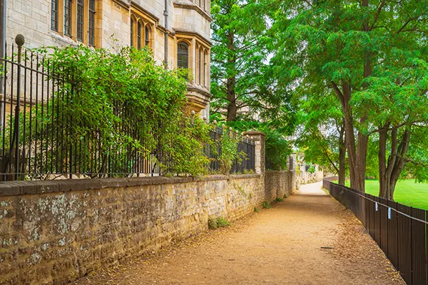 An unpaved path between a sandstone building with a brick wall and railings with plants on one side, and a park with low black railings and trees on the other. 