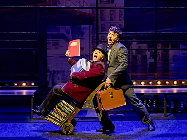 2 white actors singing on stage, one is being pushed along on a trolley while sitting on top a stack of folders. 