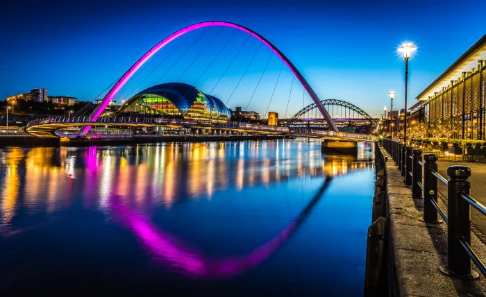 Gateshead Millennium Bridge shines in bright purple, reflected on the river below at twilight with bright lights lining the quayside walkway.