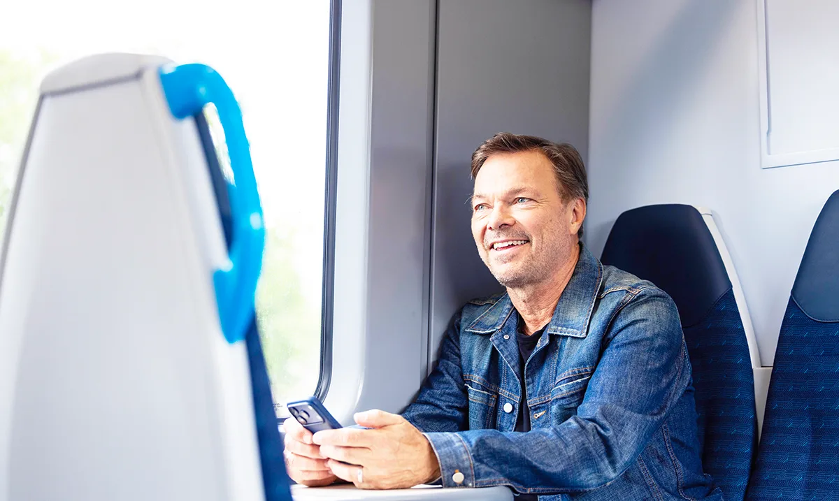 Pete Tong, a smiling middle-aged man in a denim jacket, holding a smartphone and seated by a window on a train.