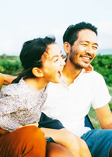 3 generations of an East Asian family – grandad, dad and daughter – are laughing and hugging each other while sitting on green grass.