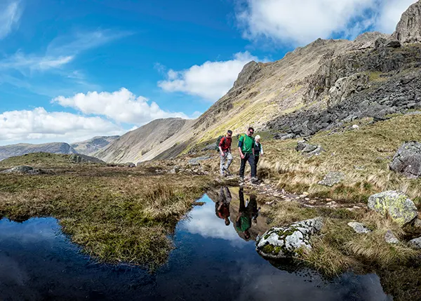 2 white men with brown hair and an older white woman with white hair, dressed in summer hiking gear and walking next to a lake in rugged, hilly countryside. 