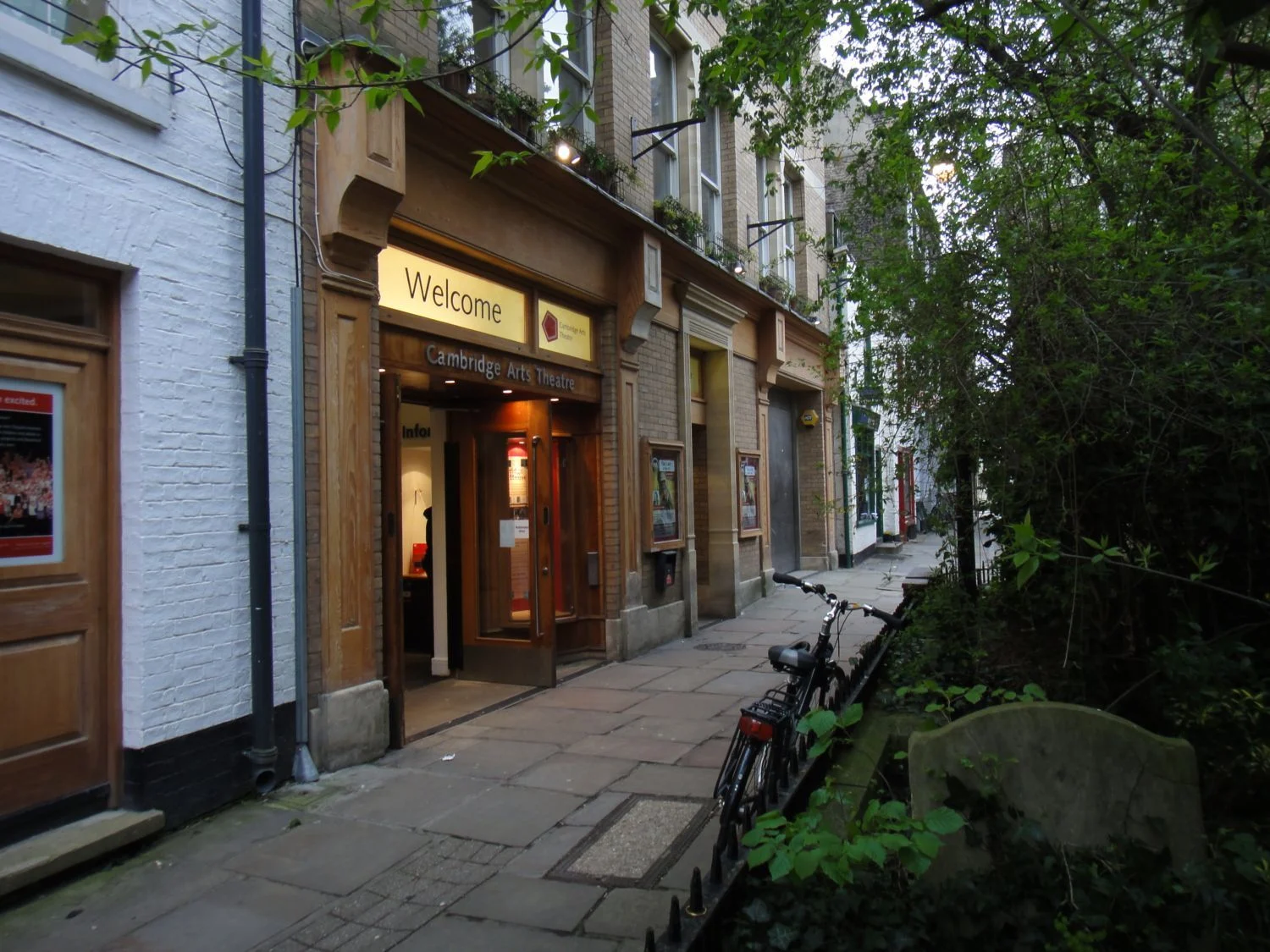 A path in a city centre, with trees on one side and buildings on the other, one of which is a small theatre.